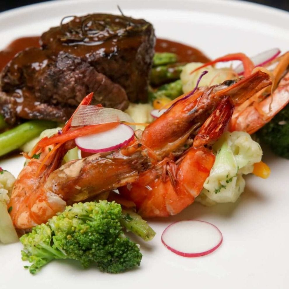 A composition of steak, shrimp, and vegetables, arranged on a clean white plate.