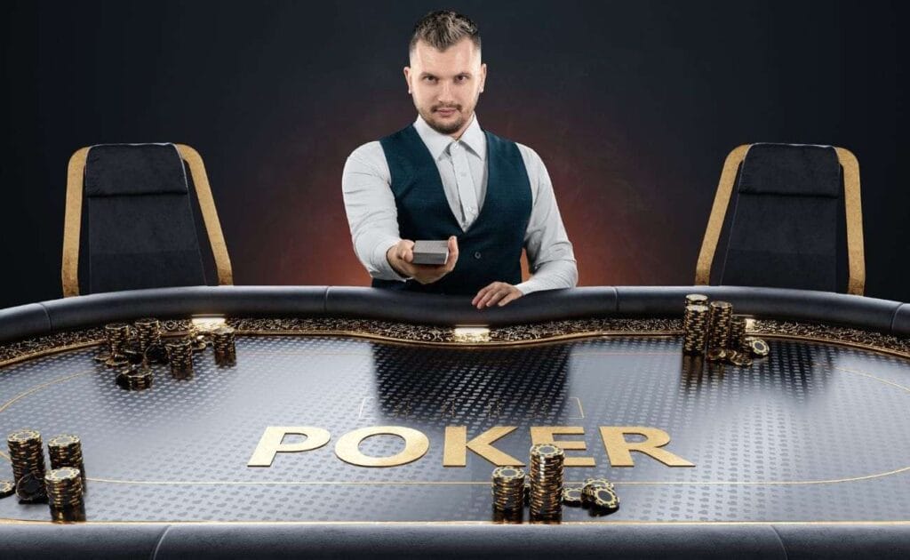 A poker dealer holds out a deck of playing cards and sits at an all-black poker table with stacks of black and gold poker chips on it.