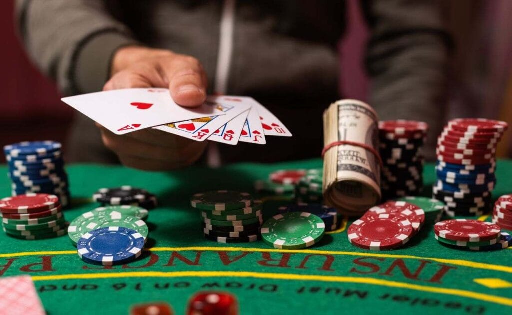 A closeup of a poker player’s hand holding a standard Royal Flush over stacks of poker chips and cash.