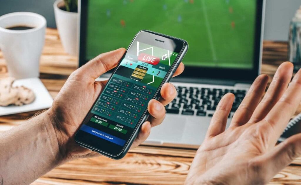 a close up of a person holding a cellphone that they are using for sports betting with a laptop in the blurred background that is playing a sports game