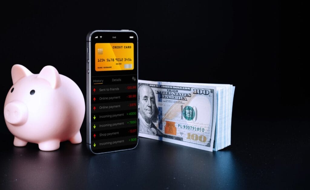 A smartphone, American dollars and piggy bank against a black background.