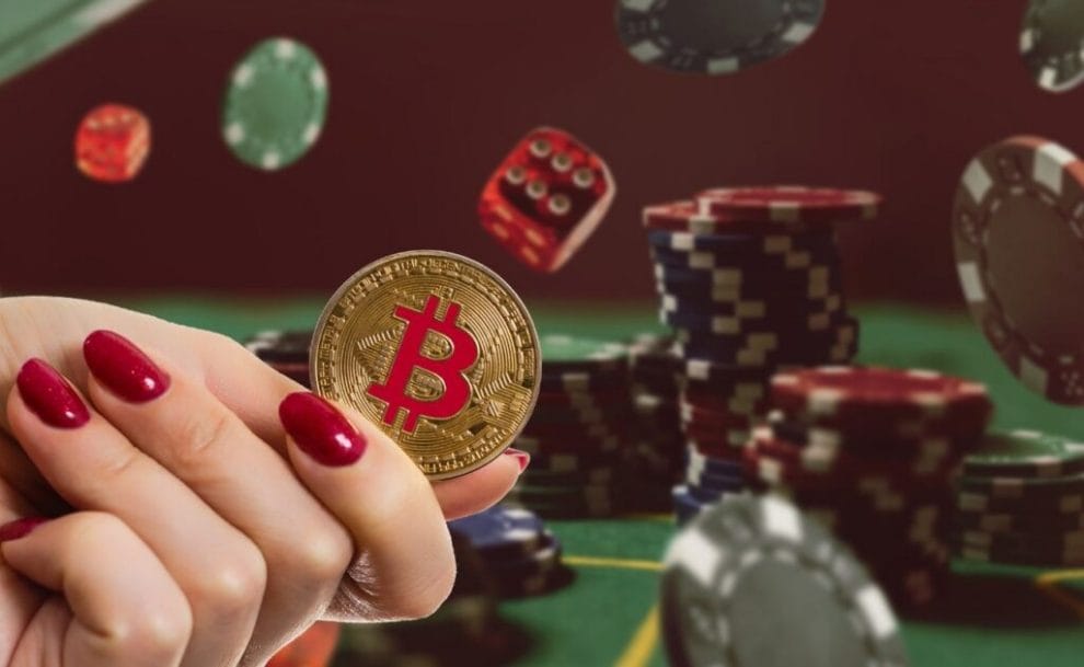 A woman's hand holding a Bitcoin coin, while in the background, a stack of poker chips rests on a poker table