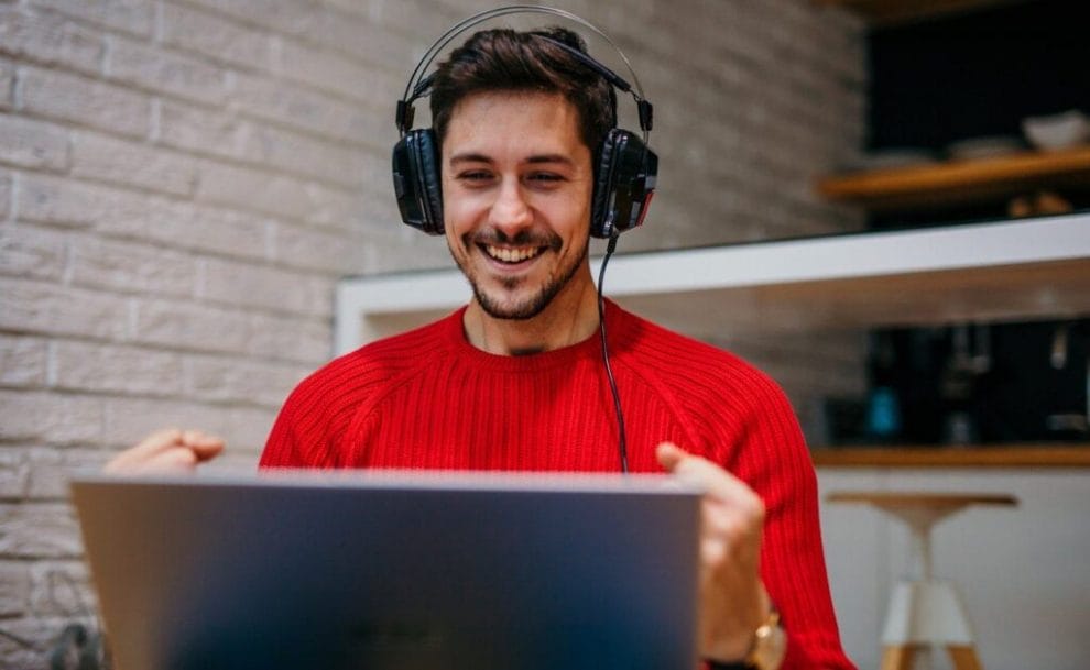 A man wearing headphones clenching his fists in victory. He is sitting in front of his laptop.