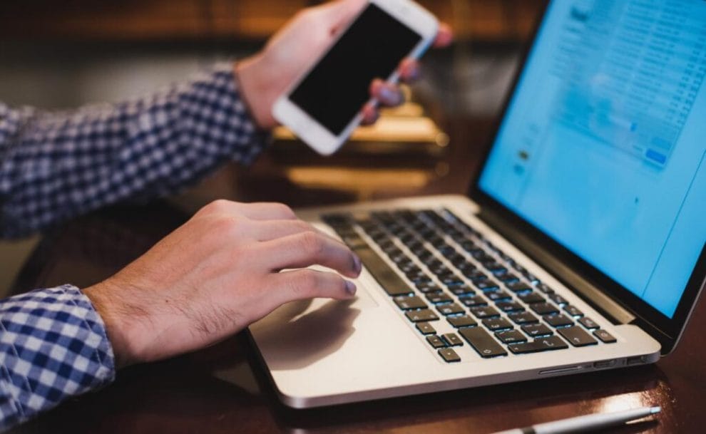 A person scrolling on a laptop with a smartphone in their hand.