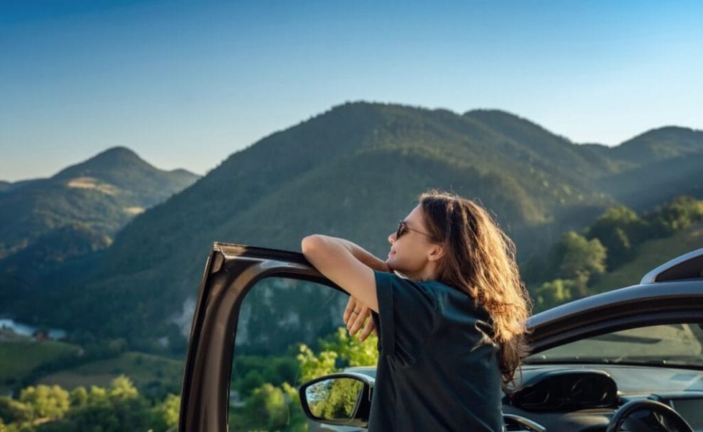 a woman standing with her arms resting on the car door as she looks out at the mountain view