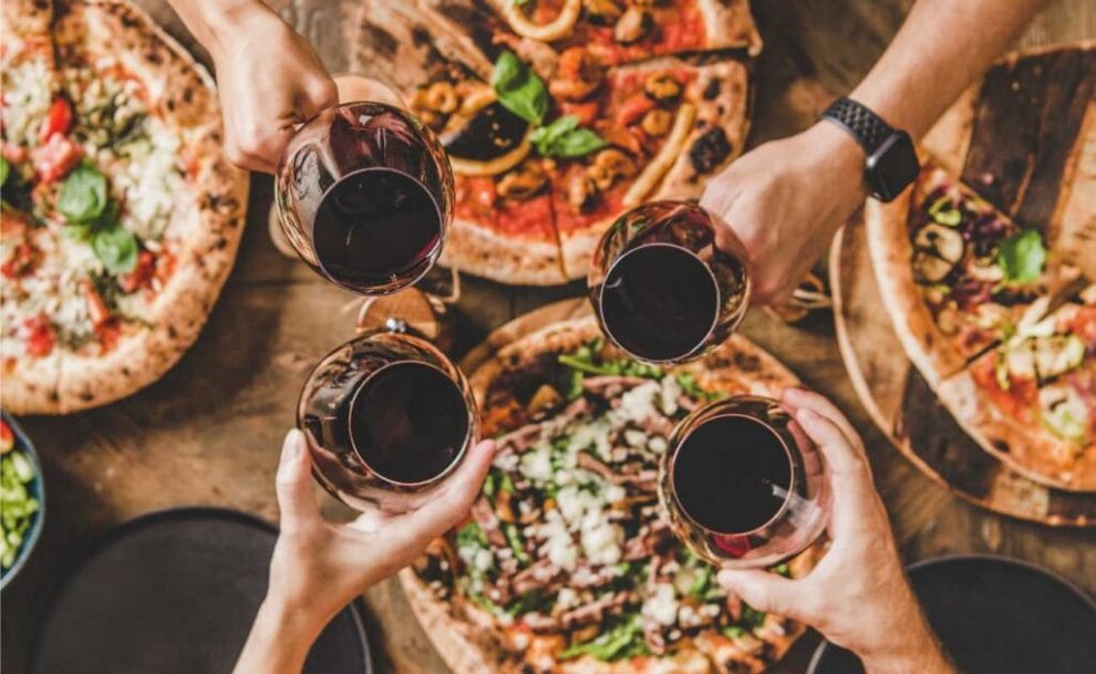 Pizzas on a table with people holding red wine glasses above.