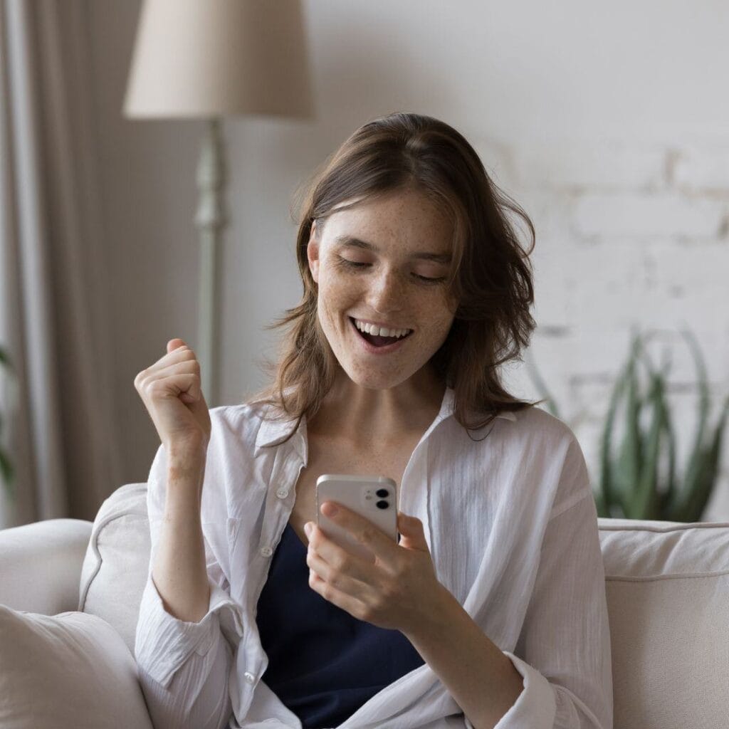 a woman is sitting on a couch and cheering with excitement while looking at the cellphone in her hand