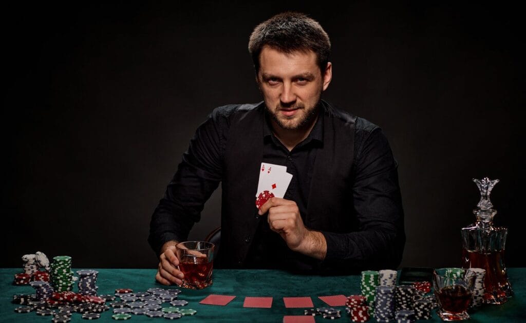 A man holding up two aces and two poker chips in the same hand. He holds his drink in the other while sitting in front of a poker table.