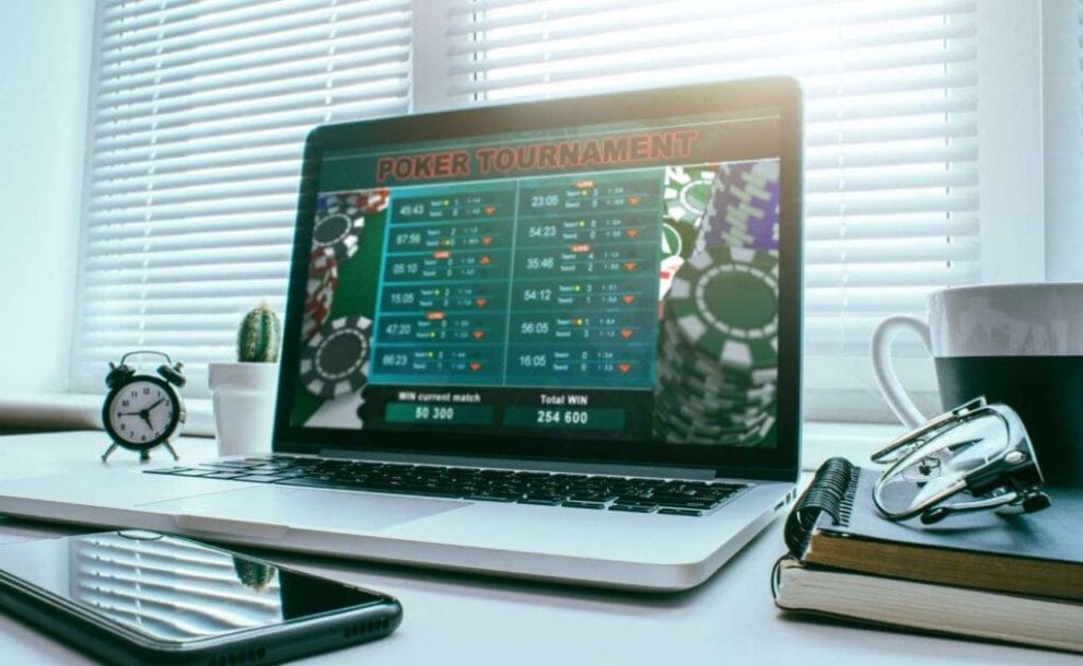 A laptop is open on a desk and displaying an online poker tournament on the screen with a cellphone, mini alarm clock, cactus pot plant, a mug, notebooks and a pair of glasses also on the desk.