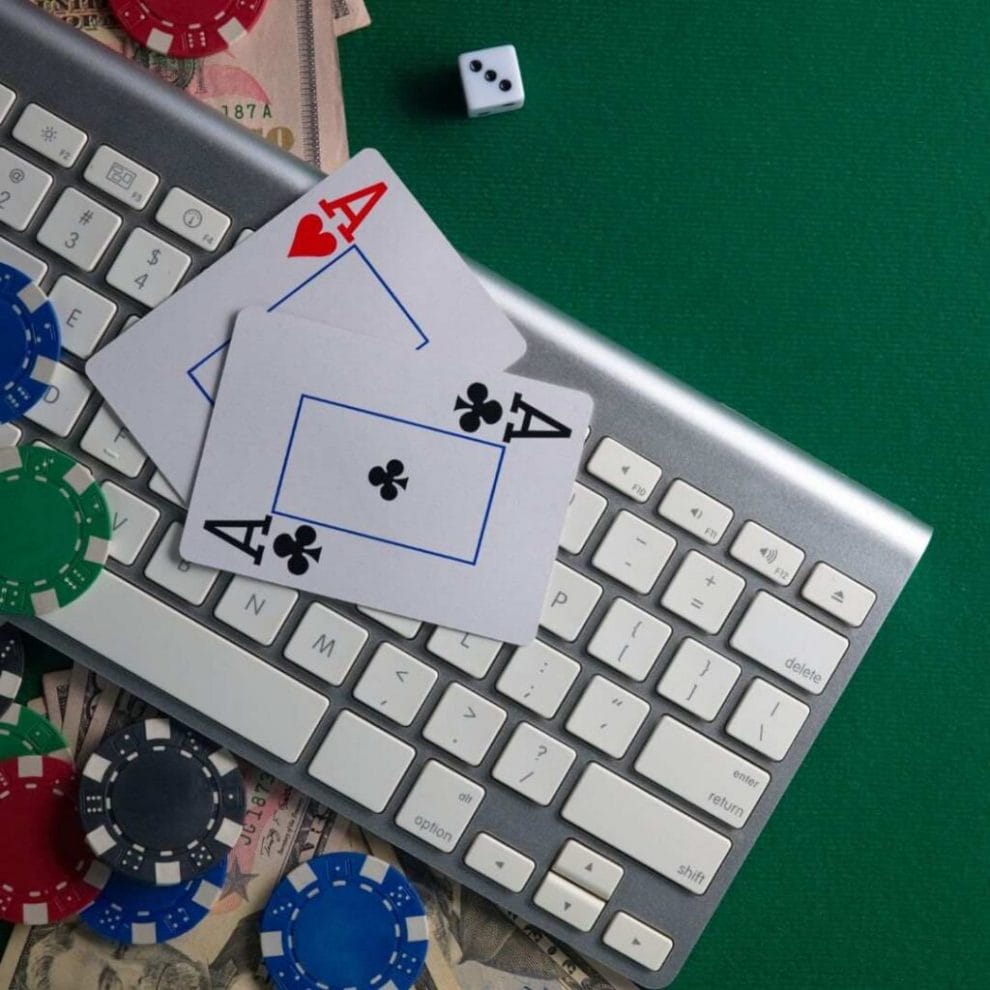 A pair of ace playing cards on a computer keyboard on a green felt surface and a white six-sided dice above it with poker chips scattered around on money bills.