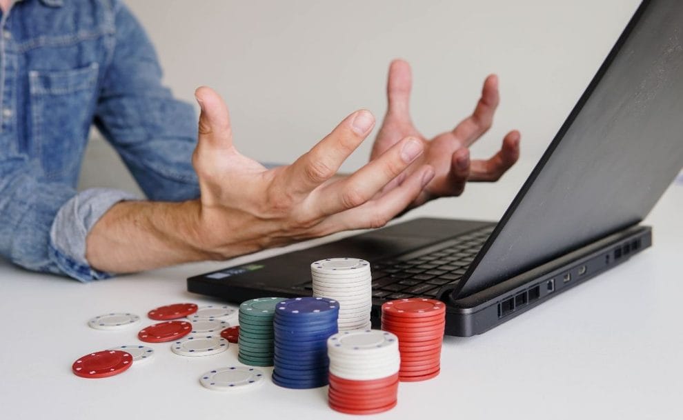 Individual displaying frustration through hand gestures while seated in front of a laptop, surrounded by poker chips on the table