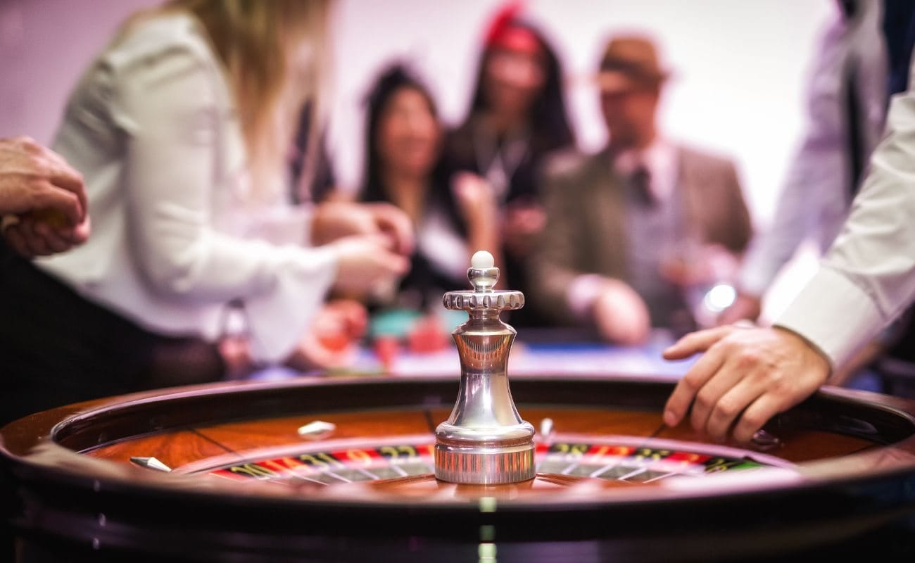 A roulette wheel in a casino, with people standing around the roulette table in the background.
