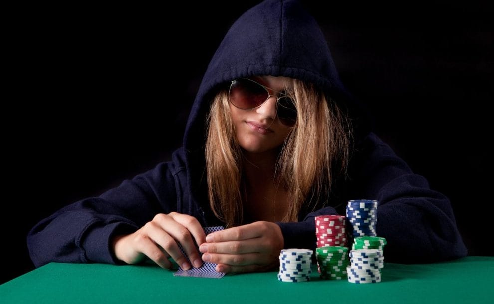 A woman wearing a hoodie and sunglasses holding playing cards in her hands, seated at a poker table, with stacks of casino chips in front of her.