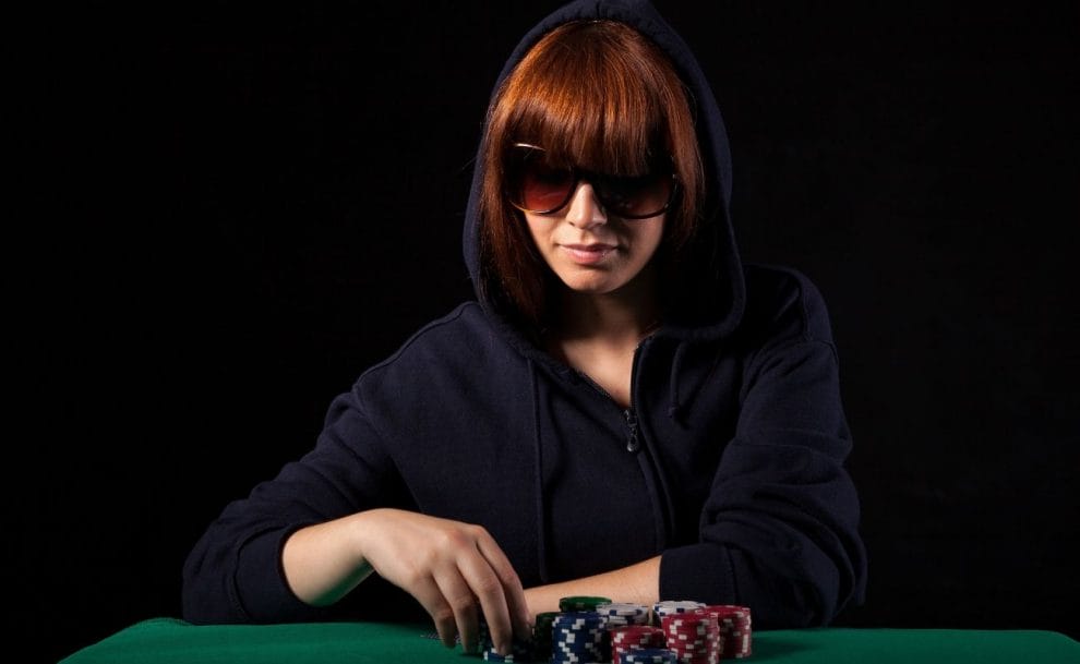 A woman wearing a hoodie and sunglasses, sitting at a poker table, with stacks of poker chips placed in front of her.