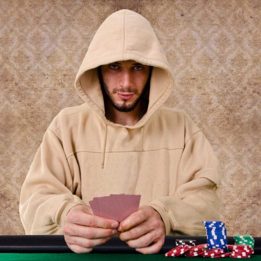 A man wearing a hoodie holding playing cards in his hands, seated at a poker table, with a few stacks of casino chips in front of him.