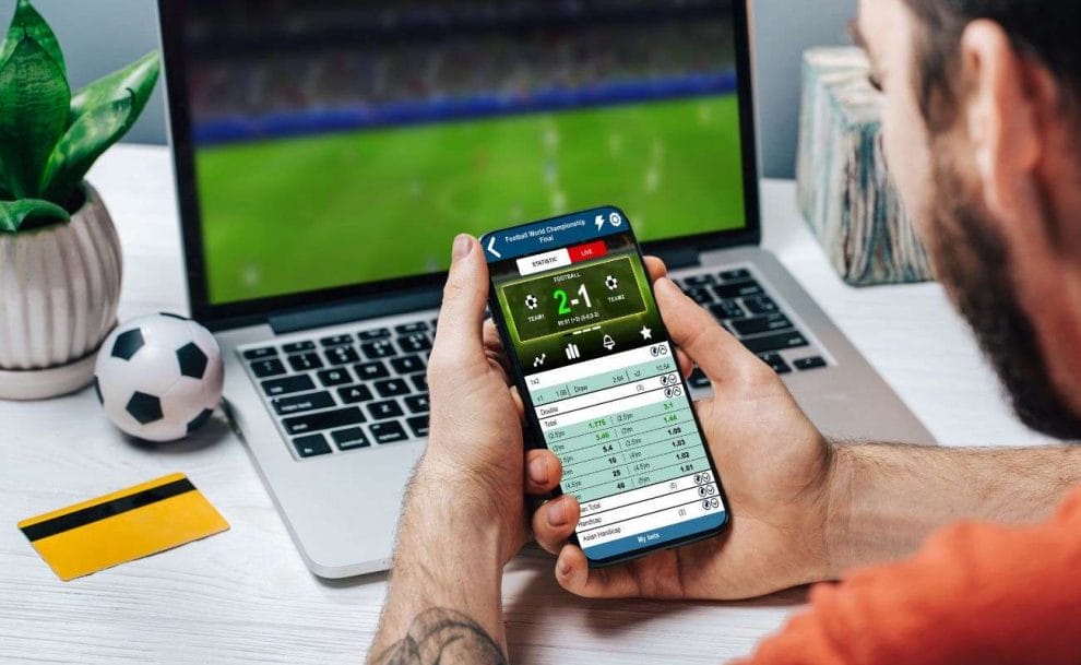 An over-the-shoulder view of a man sitting at his laptop with his cellphone in his hand on a sports betting website, and a small soccer ball and his bank card beside him.
