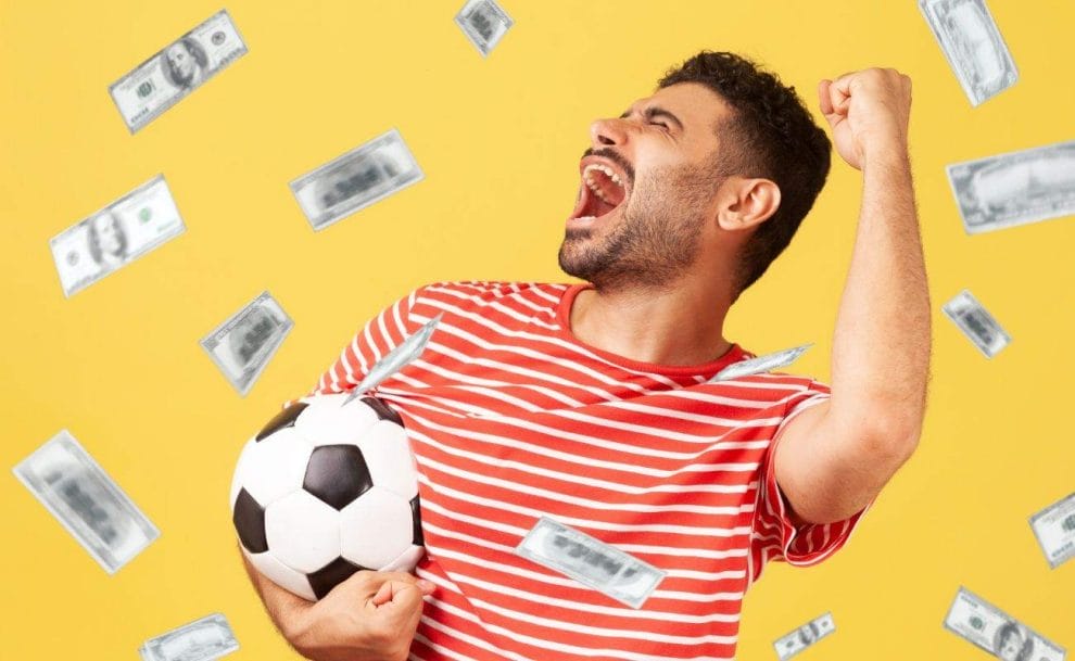 A cheering man in a red and white shirt stands in front of a yellow background holding a soccer ball with cash floating around him. 