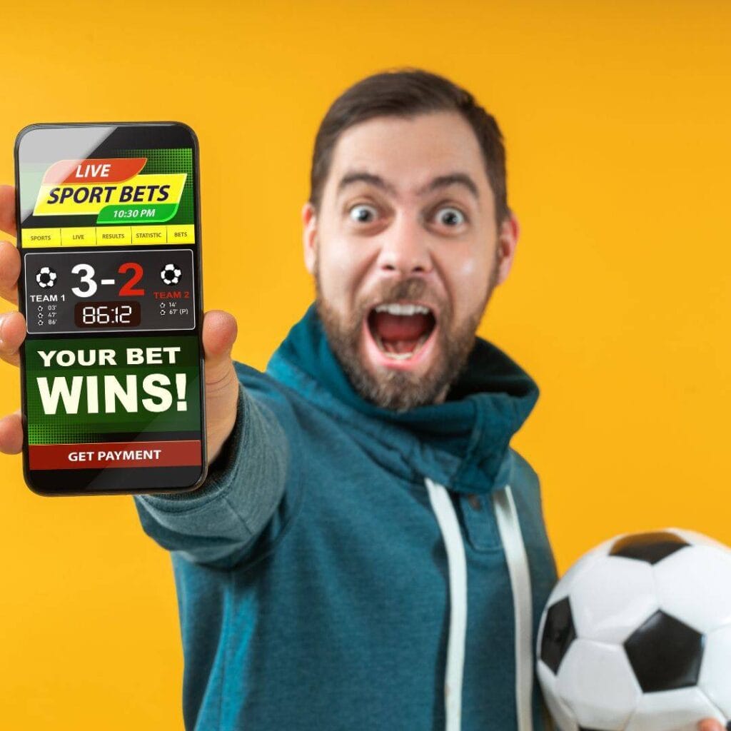 A very excited man stands in front of a yellow background holding a soccer ball and shows the camera his winning sports bet on his cellphone.