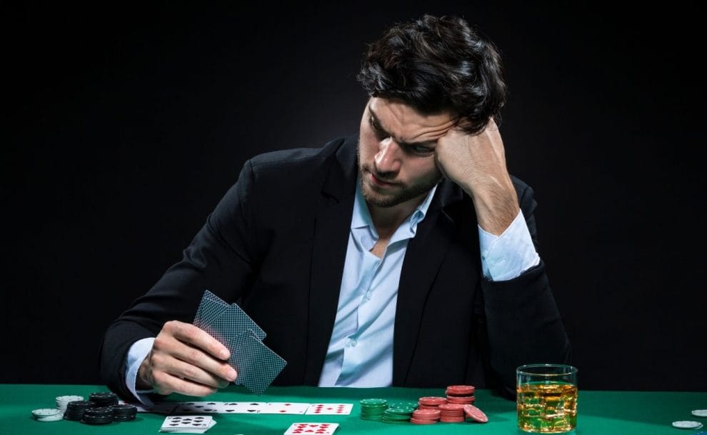 a frustrated man sits at a green felt poker table, resting his head on his fist as he examines the playing cards in his hands, the table is scattered with poker chips, playing cards, and a beverage