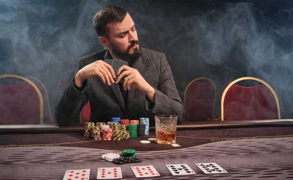 poker player seated at a table with cards, chips, and a drink, holding two cards, with smoke in the air