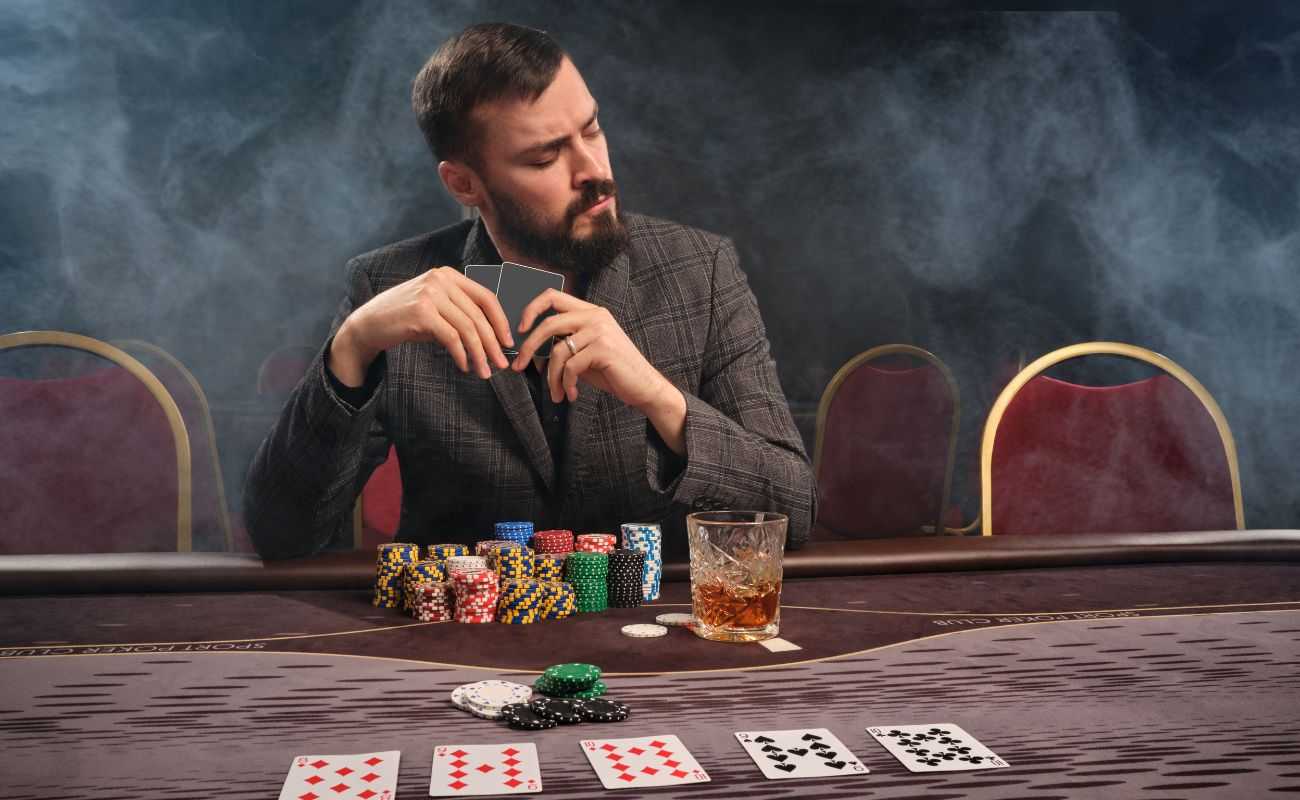 poker player seated at a table with cards, chips, and a drink, holding two cards, with smoke in the air