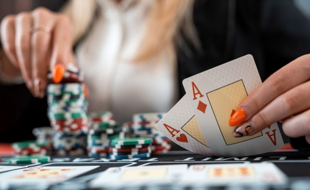 A female poker player holds up two Ace playing cards with one hands while removing chips from her stack with the other to place a bet. 