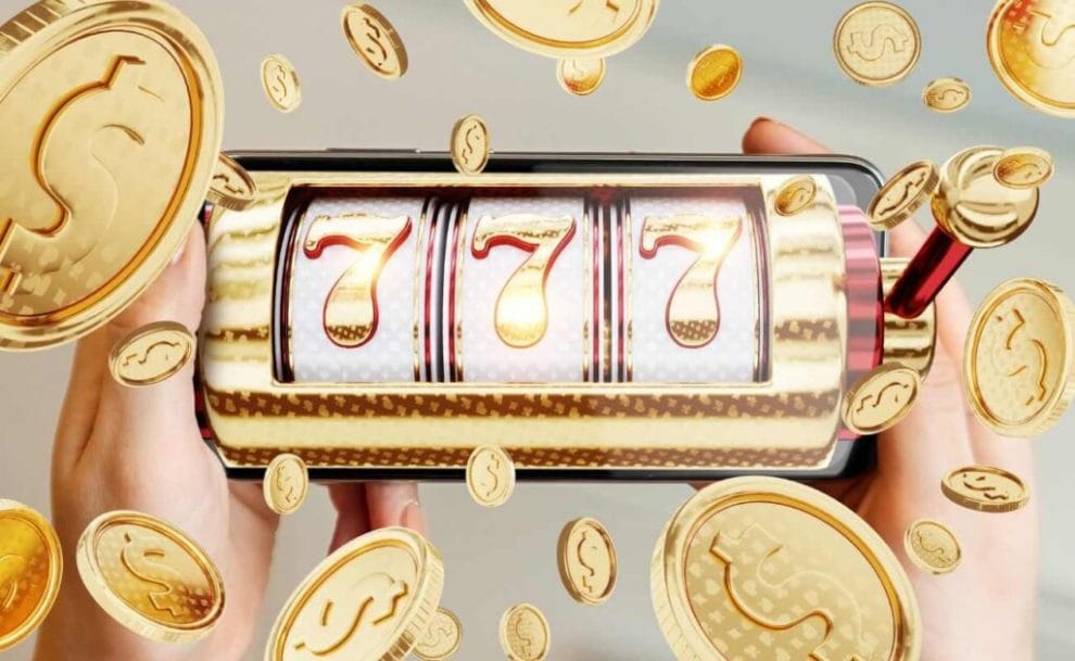 A closeip of a cellphone in a woman’s hands with a gold slot reel showing three 7s on the screen and gold coins floating around the image.