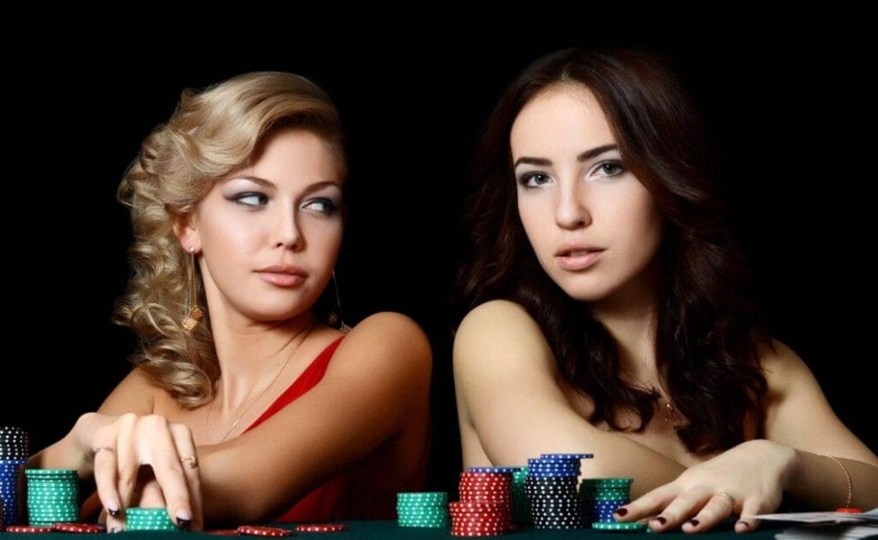 Two beautiful women sit at a table with stacks of poker chips in front of them infront of a black background.