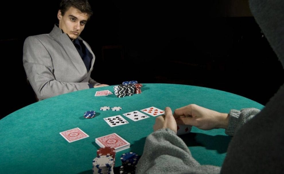 Two men dressed in grey sit at a round table and play a game of poker. 