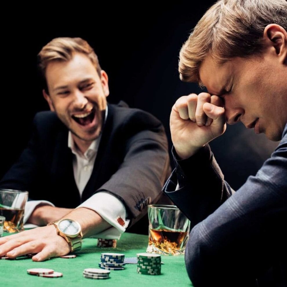 Two friends sit at a poker table, one with his hand on his head in frustration and the other laughing as he reaches for the poker chips.