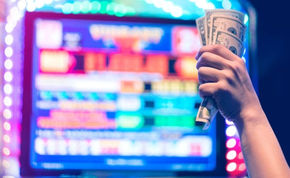 A hand holding money in front of a slot machine.