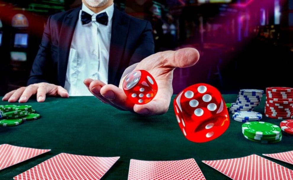 A dealer throws two red six-sided dice on a green felt table with five face-down cards on it and stacks of casino chips on each side of the image.