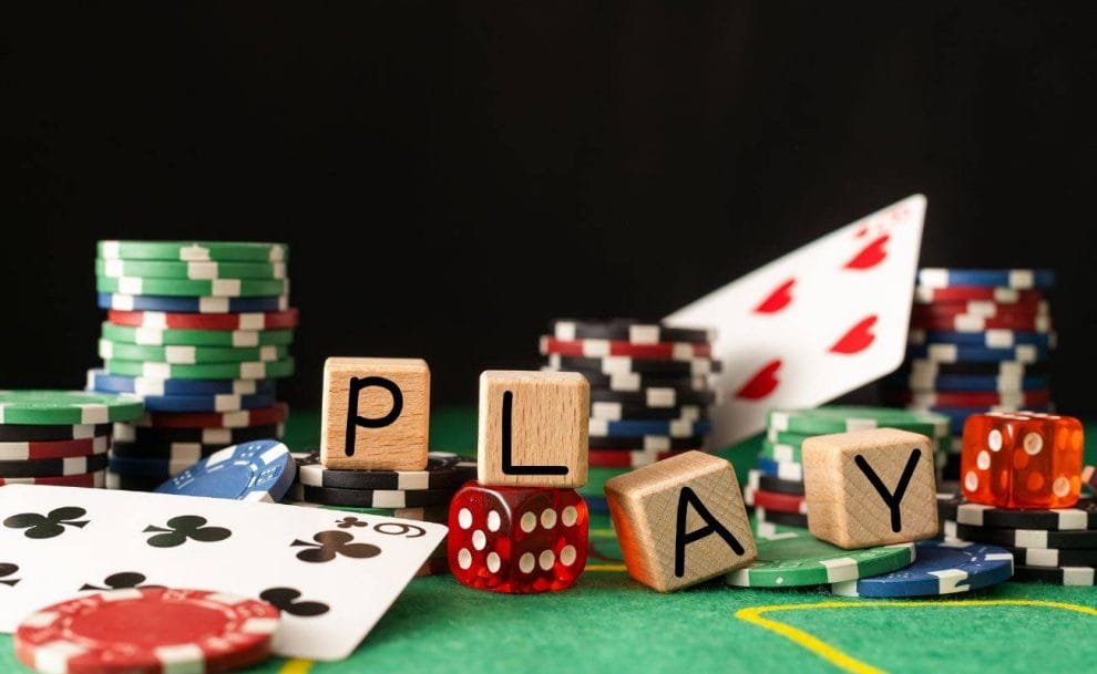 Four wooden blocks spelling out the word “play” balancing on top of poker chips and dice on a green felt table with two playing cards in between the chips.