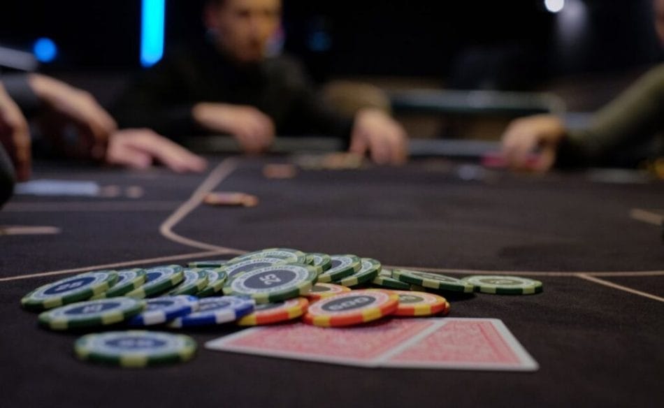 poker chips scattered on a poker table next to two playing cards during a poker game with the other players sitting around the table in the blurred background.