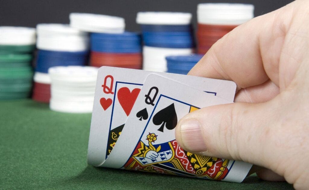 a person checking their hole cards of a pocket pair of queens on a green felt poker table with stacks of poker chips blurred in the background
