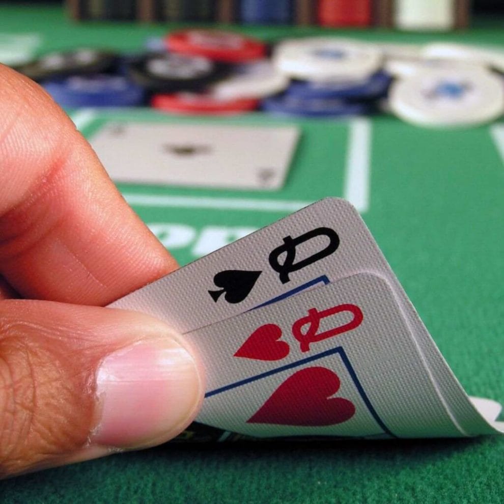 a person checking their hole cards of a pocket pair of queens on a green felt poker table during a game of poker with poker chips blurred in the background