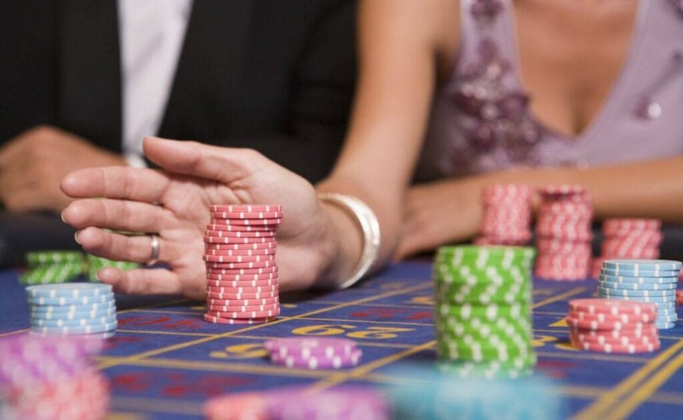 a woman sitting next to a man pushing a stack of poker chips on a roulette table with other poker chip stacks on it in a casino 