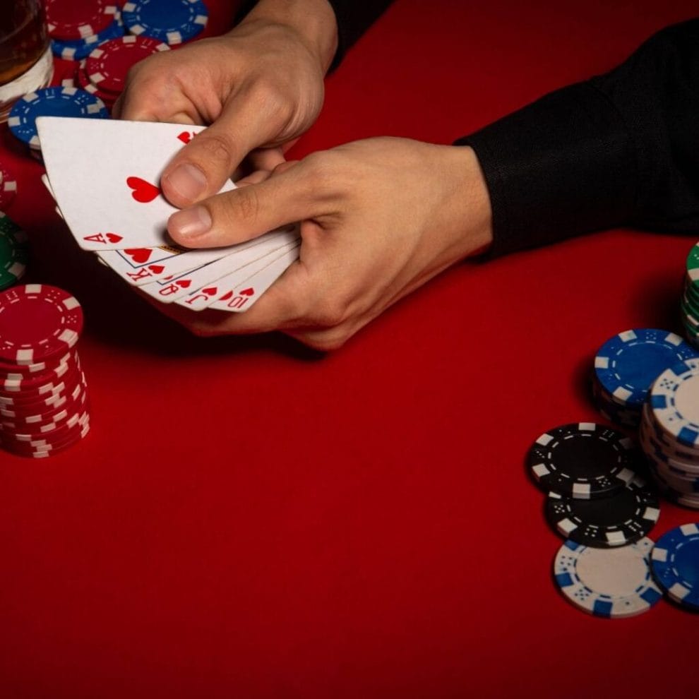 A poker player holding his cards over a red felt table with stacks of poker chips on either side of his arms.