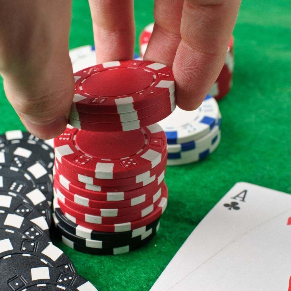 A closeup of a hand taking a few poker chips off the top of a small chip stack next to two Ace playing cards on a green felt table.