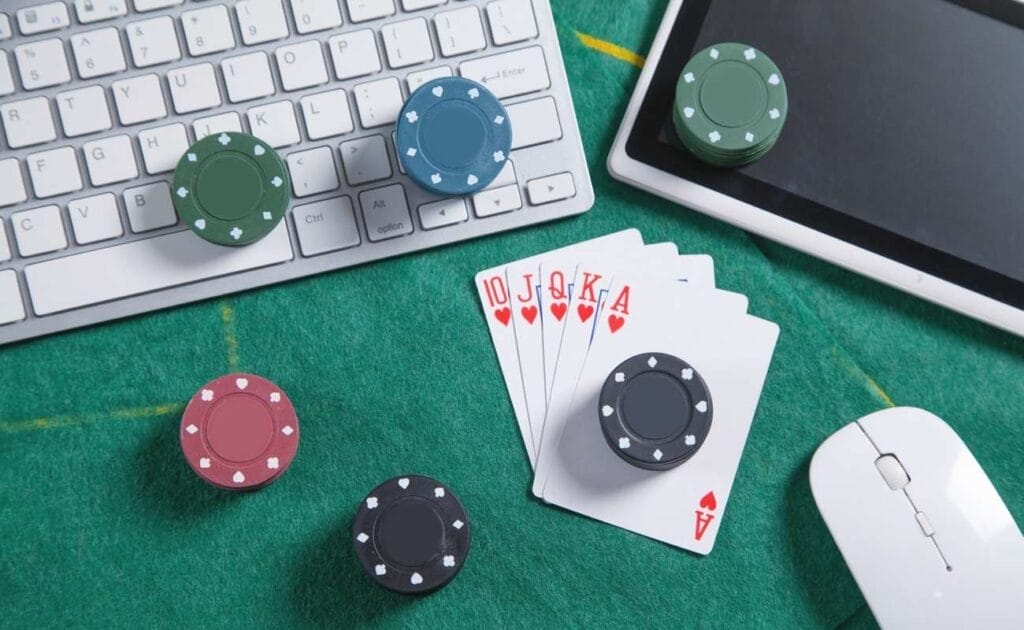 A keyboard, an Ipad, a mouse, 6 stacks of poker chips, and 10, jack, queen, king, and ace of hearts on a green felt poker table.