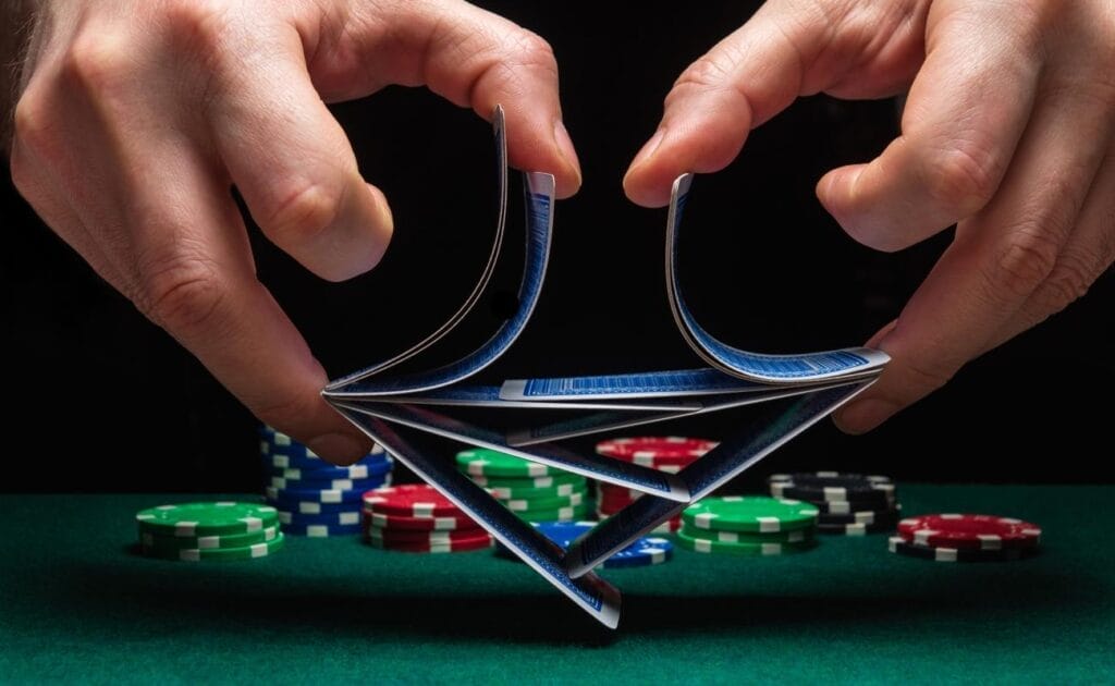 a pair of hands shuffles a deck of cards with stacks of poker chips in the background, on a green felt poker table.