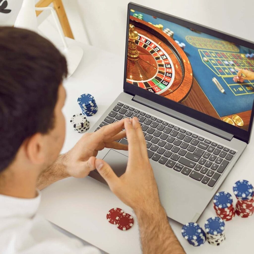 A man sits in front of a laptop, with online casino gameplay on the screen and stacks of poker chips either side of the keyboard