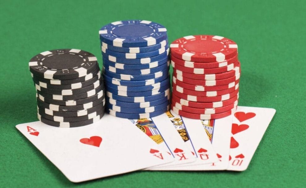 An ace, king, queen, jack and ten of hearts are fanned out, lying face up on a green felt poker table with stacks of poker chips laying on top of them.