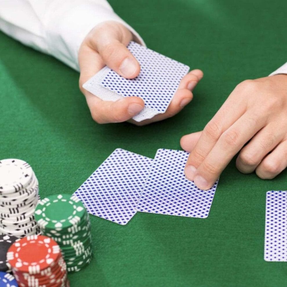 Live dealer is holding a deck of cards and is dealing two playing cards on a green felt table, with stacks of poker chips on the left.