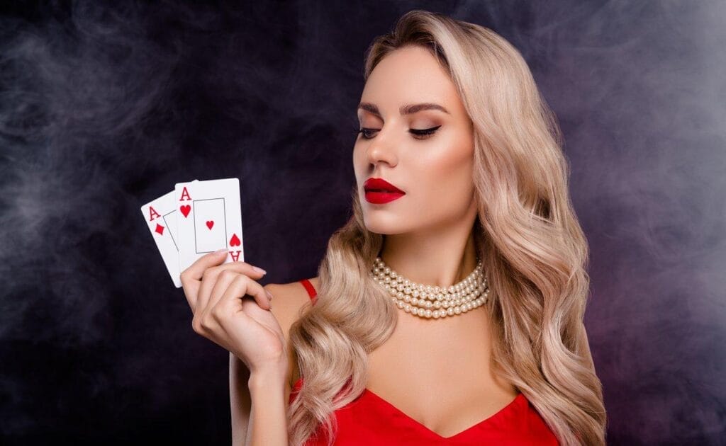 A person in a red dress and pearls holds up two aces.