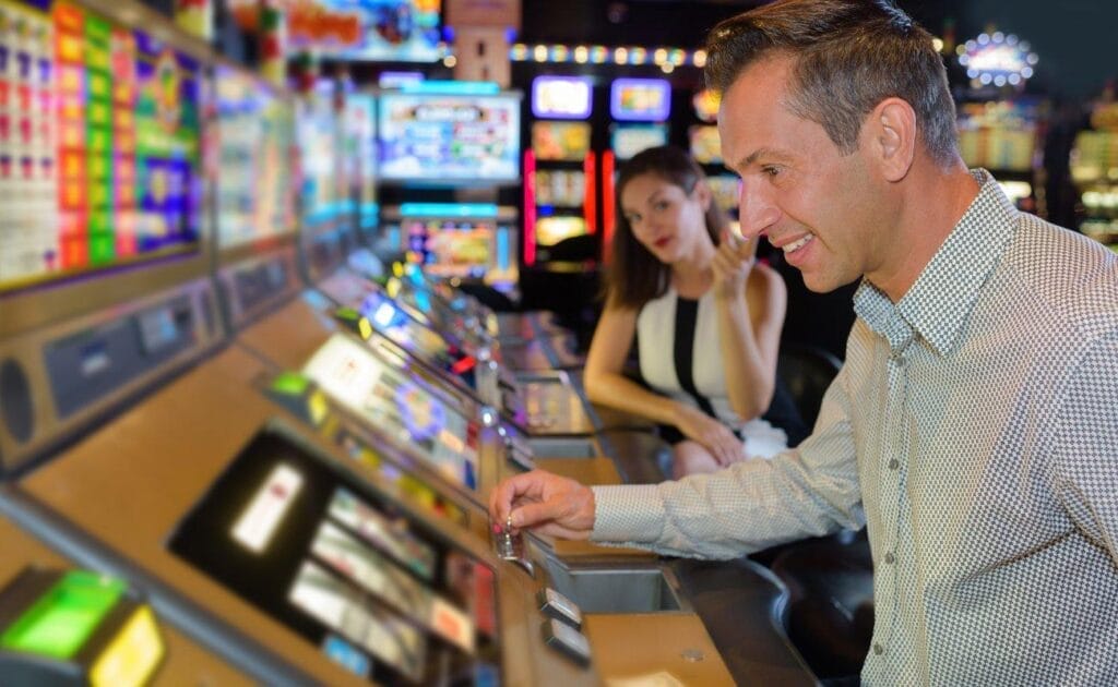 A person playing slot machines in a casino while another person in the background looks on.