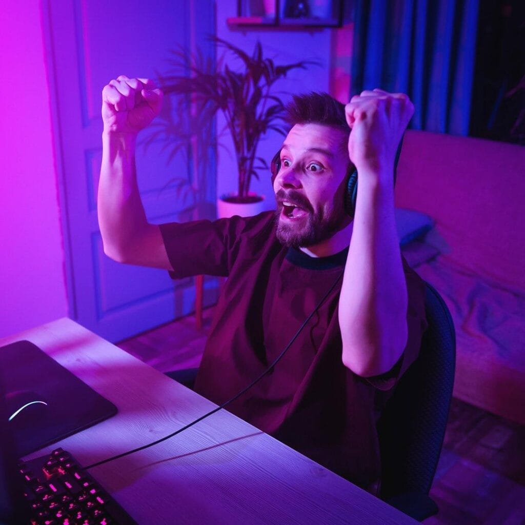 A surprised and happy person wearing headphones in front of their computer. They raise their hands in victory.