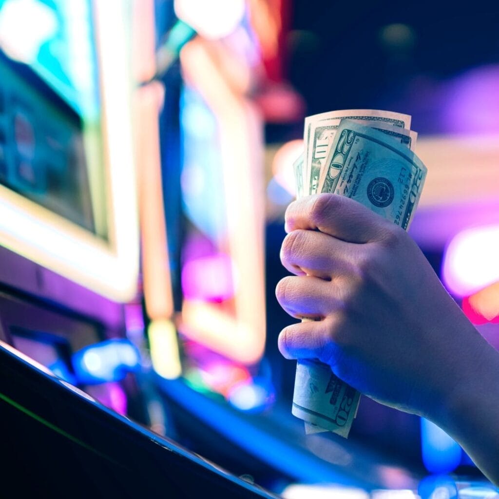 A hand holds a multiple dollar bills in front of a slot machine
