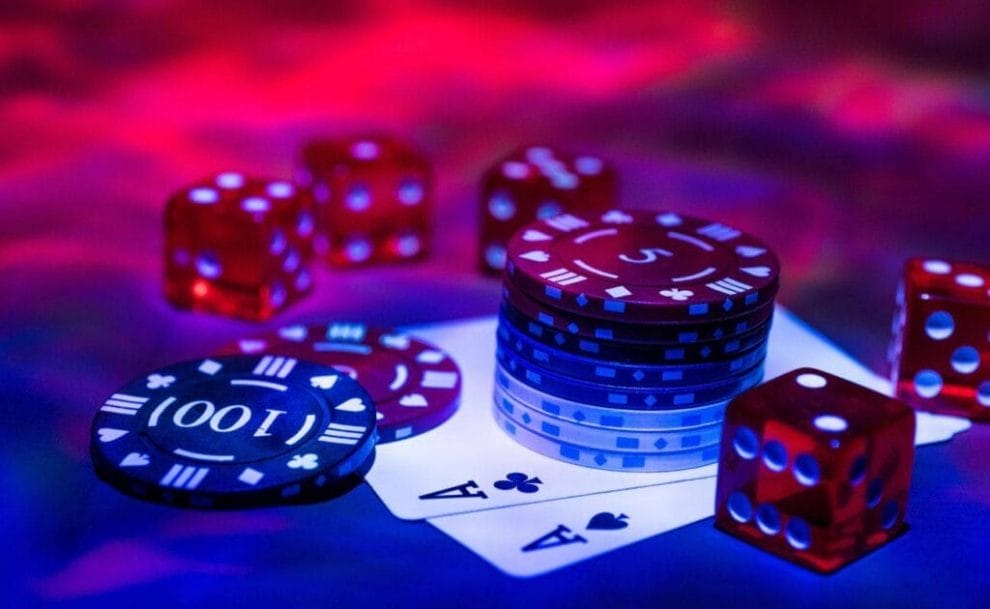 Poker chips and five red six sided dice on top of two Ace playing cards in blue and red ambient lighting.