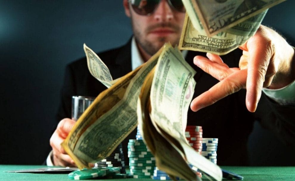 A man in a suit sits at a poker table with a drink in his and and stacks of poker chips in front of him and throws money towards the camera. 